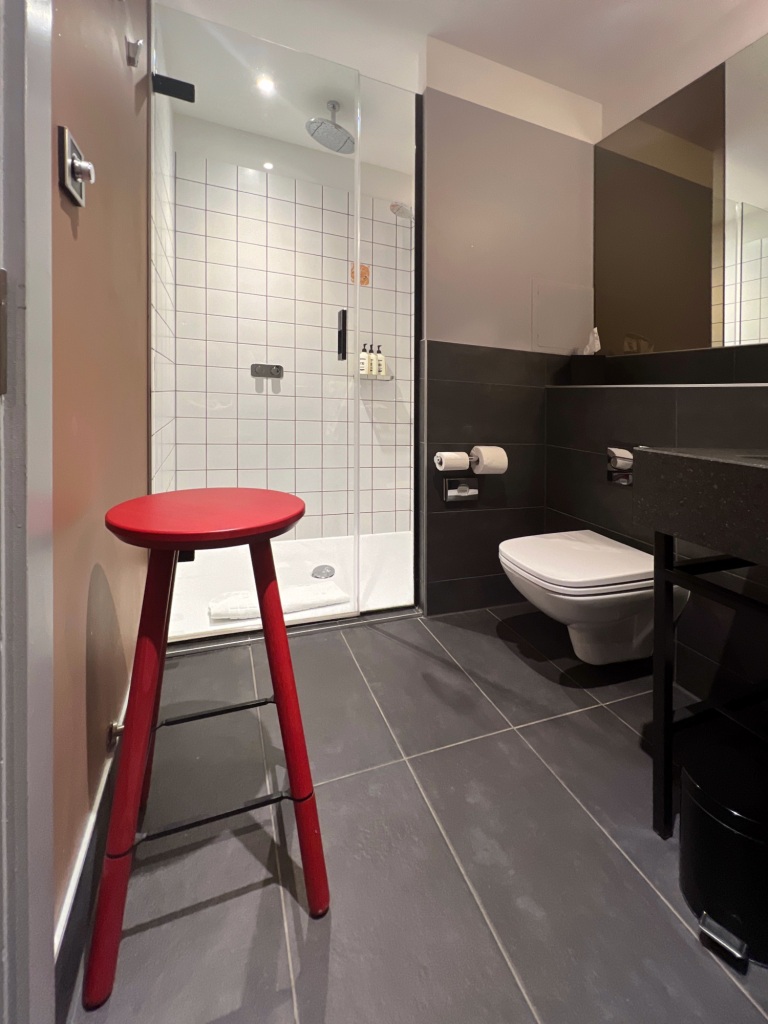 A bathroom with a bright red stool on the left, a shower in the background, and a cludgie and sink on the right.