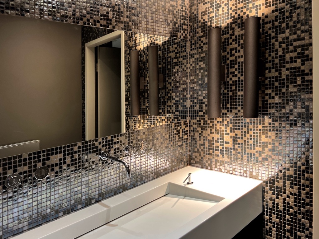 A view of a long white sink with soft lights to the right, and a mirror on the wall behind the sink. The walls look as if they are made of many small tiles in different brown shades.