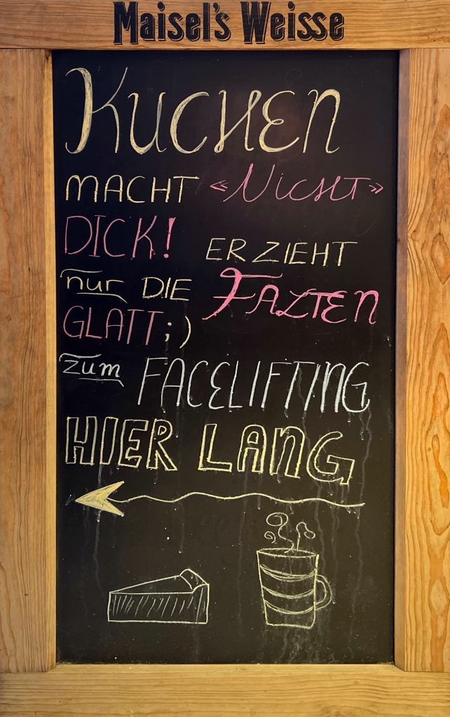 A chalkboard with the following written on it: “Kuchen macht nicht dick! Er zieht nur die Falten glatt ;) Zum Facelifting hier lang” and then with an arrow pointing left, and a sketch of a cake and a coffee below.