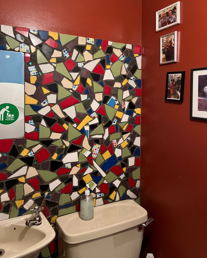 A colourful tiled wall on the left above a sink and a cludgie, and on the right a red wall with some small framed pictures.