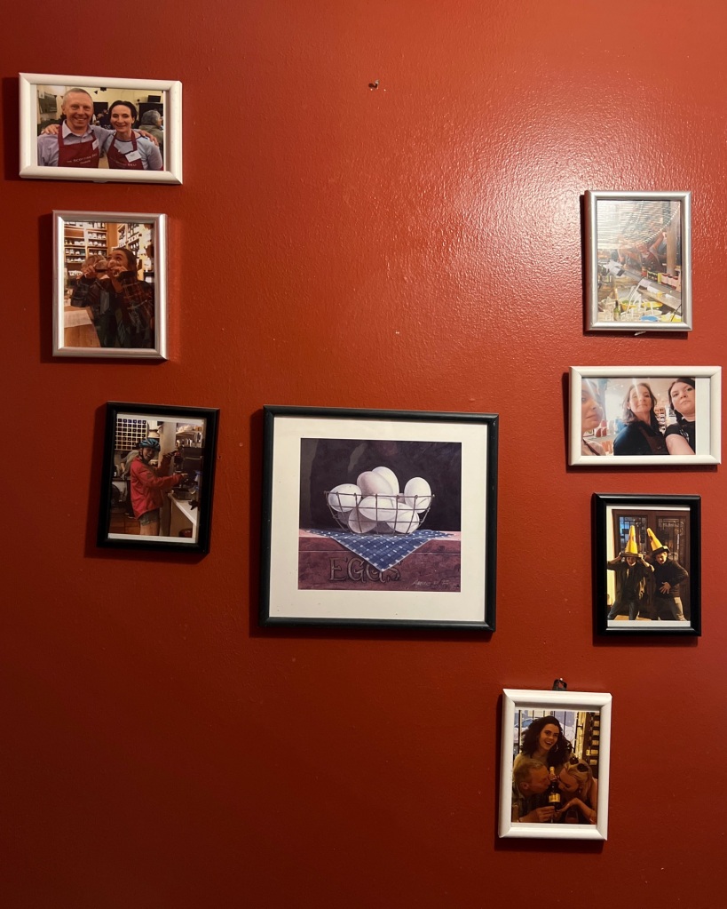 Six small framed photographs on a wall, of various people, with a painting in the middle of several eggs in a basket. The wall is a deep dark read.