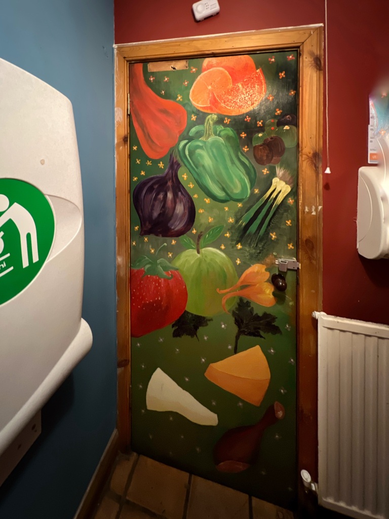 A colourful door, covered in pictures of fruit, vegetables and cheeses. On the right of the door there is a radiator, and on the left a fold-out baby-changing table.